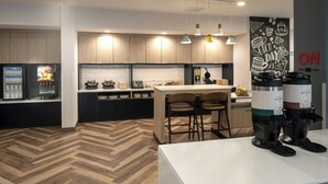 A modern kitchen with wooden cabinets, a herringbone floor, a central island with bar stools, a coffee machine, and a wall-mounted sign. - TownePlace Suites by Marriott West Lafayette (West Lafayette)