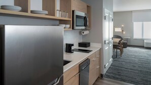 A modern kitchen with a stainless steel refrigerator, wooden cabinets, and a microwave. There is a countertop with a coffee maker and a dishwasher. The kitchen leads to a dining area with a table and chairs. - TownePlace Suites by Marriott West Lafayette (West Lafayette)