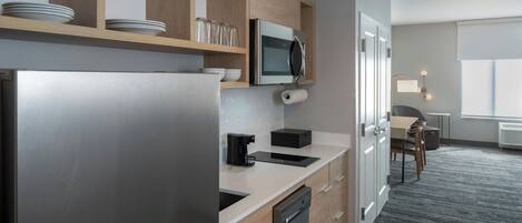 Cocina moderna con refrigerador de acero inoxidable, armarios de madera y microondas. Hay una encimera con cafetera y lavavajillas. La cocina da a un comedor con mesa y sillas.
