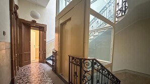 Un pasillo con piso estampado, una puerta de madera, una escalera con barandillas de hierro forjado y un ventanal con vistas a edificios.