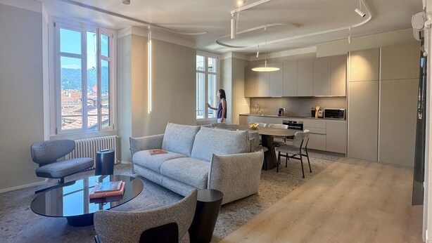 Una sala de estar moderna con un sofá, una mesa de centro y un comedor con sillas. Hay una zona de cocina con armarios y una ventana con vistas a un paisaje urbano.