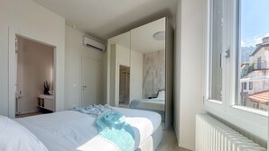Un dormitorio con cama, espejo, radiador y ventana con vistas a edificios.