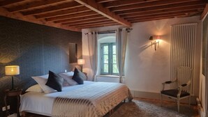 Un dormitorio con techo de madera, una cama con ropa de cama blanca, una mesita de noche con lámpara, una silla y una ventana con cortinas.