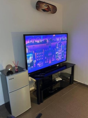 A flat-screen TV on a black stand with a cityscape wallpaper, a white mini-fridge, and a small decorative item on top. - Acogedor Apartamento en el Corazón de la CDMX – SúperAnfitrión Coyoacán (Ciudad de México)