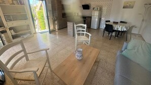 A living room with a wooden coffee table, white chairs, a grey sofa, a kitchen area with a TV, and a bookshelf. - Superb sea view apartment with private pool! 5 minutes from the center of Roses. (Roses)
