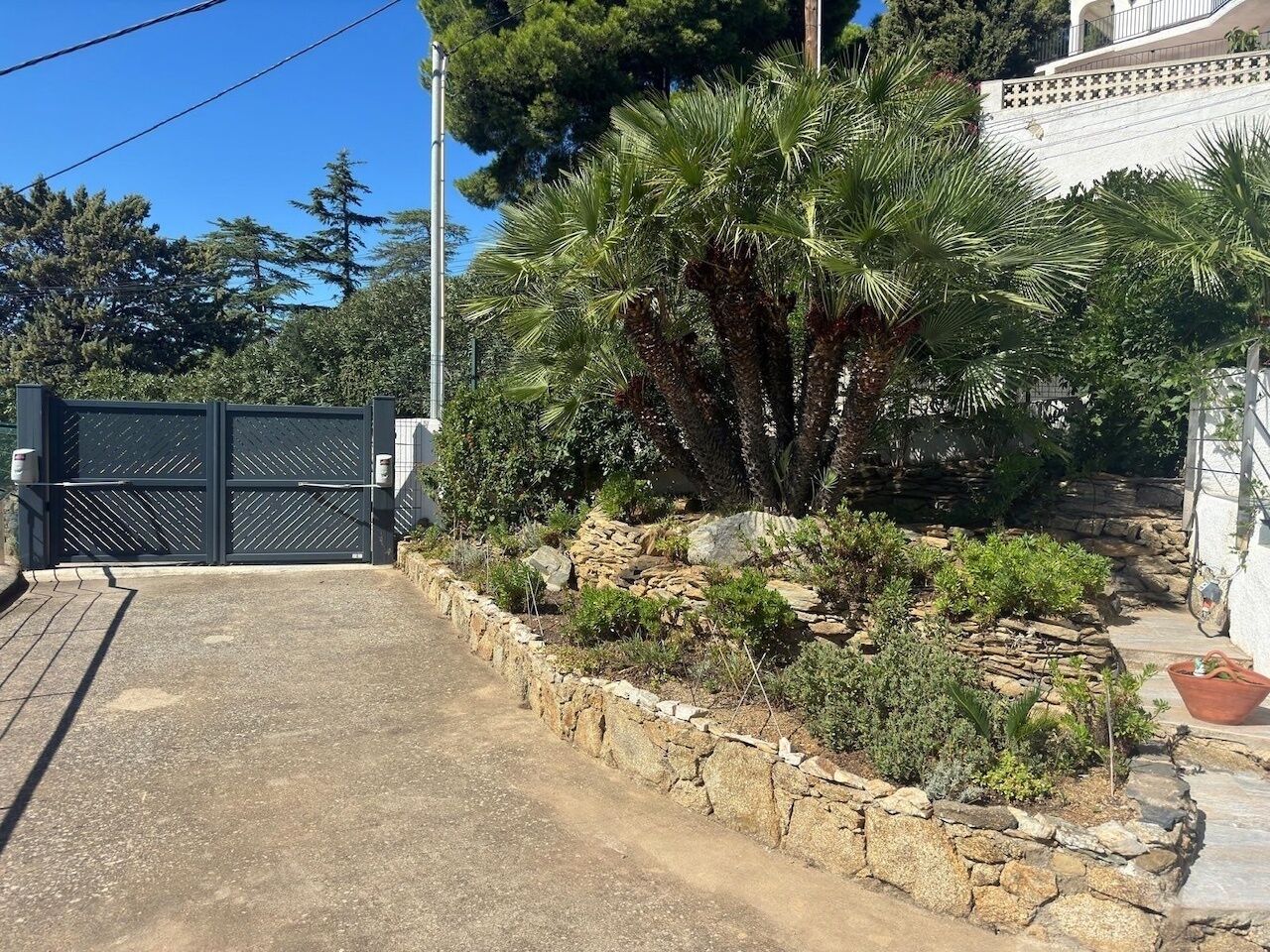 Une allée privée fermée par un portail, avec un mur de soutènement en pierre, des palmiers et un bâtiment blanc à la façade recouverte de tuiles.