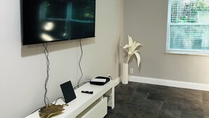 Una sala de estar moderna con un televisor de pantalla plana montado en la pared, un mueble blanco para entretenimiento con una computadora portátil y un control remoto, un ventanal con persianas y una alfombra estampada sobre el suelo oscuro.