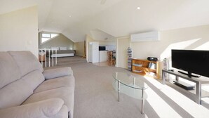 Un vaste espace de vie comprenant un canapé, une table basse en verre, un téléviseur posé sur un meuble et un ventilateur de plafond.