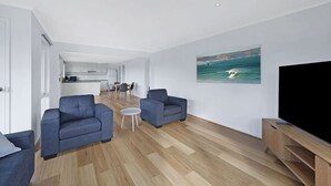 Een moderne woonkamer met twee banken, een salontafel en een flatscreen televisie. Achtergrond is een eethoek, met zicht op een keuken.