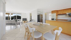 Une cuisine moderne avec des meubles en bois, un coin repas avec une table en bois et des chaises blanches, et une vue sur l’extérieur grâce à de grandes fenêtres.
