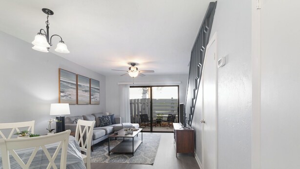 A modern living room with a sofa, coffee table, and a dining area with a chair and table. There is a ceiling fan and a view of the outdoors through a sliding glass door. - Modern & Stylish Condo Near UF - Foxmoor (Gainesville)