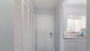 A narrow hallway leading to a white door, with a kitchen area on the right featuring a window, sink, and dish soap. - Modern & Stylish Condo Near UF - Foxmoor (Gainesville)