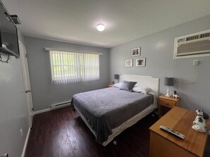 Een slaapkamer met een bed, een nachtkastje, een lamp, een afstandsbediening en een wandairconditioner.