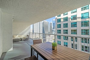Un balcon dotĂ© dâune table en bois et dâun espace salon agrĂ©mentĂ© de coussins, offrant une vue sur un paysage urbain avec des bĂątiments modernes.