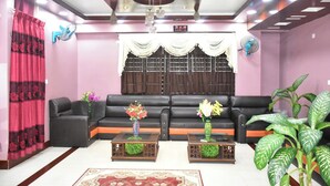 A living room with a black sofa, two side tables with vases, a large window with curtains, a ceiling fan, and a potted plant. - Hotel Aranna Residential Bandarban (Bandarban)
