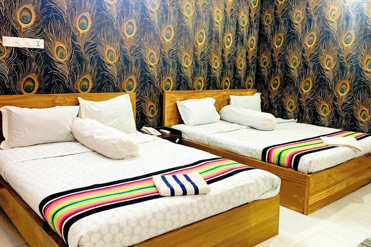 Deux lits en bois, recouverts de draps blancs et de couvertures rayées aux couleurs vives, sont placés contre un mur orné d’un motif de plumes de paon.