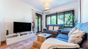 A modern living room with a flat-screen TV, a grey sofa, a coffee table, and a large window offering a view of greenery. - CT 349 - Stupa Hills - Benalmadena (Benalmádena)