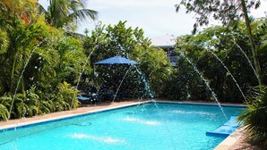 En swimmingpool med vattenfontäner, omgiven av frodig grönska och ett blått parasoll.