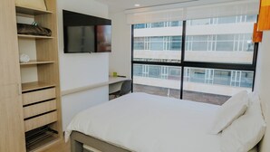 A bedroom with a bed, a TV mounted on the wall, a window with a view of a building, and a wooden wardrobe with shelves.