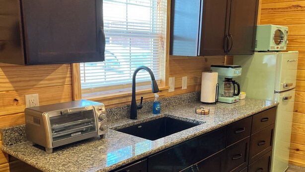 Uma cozinha compacta com armários escuros, bancada de granito, torradeira embutida, cafeteira e geladeira.