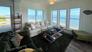 Un salón con un sofá blanco, un sillón verde y una mesa de centro con un rompecabezas. Amplias ventanas ofrecen vistas al mar.