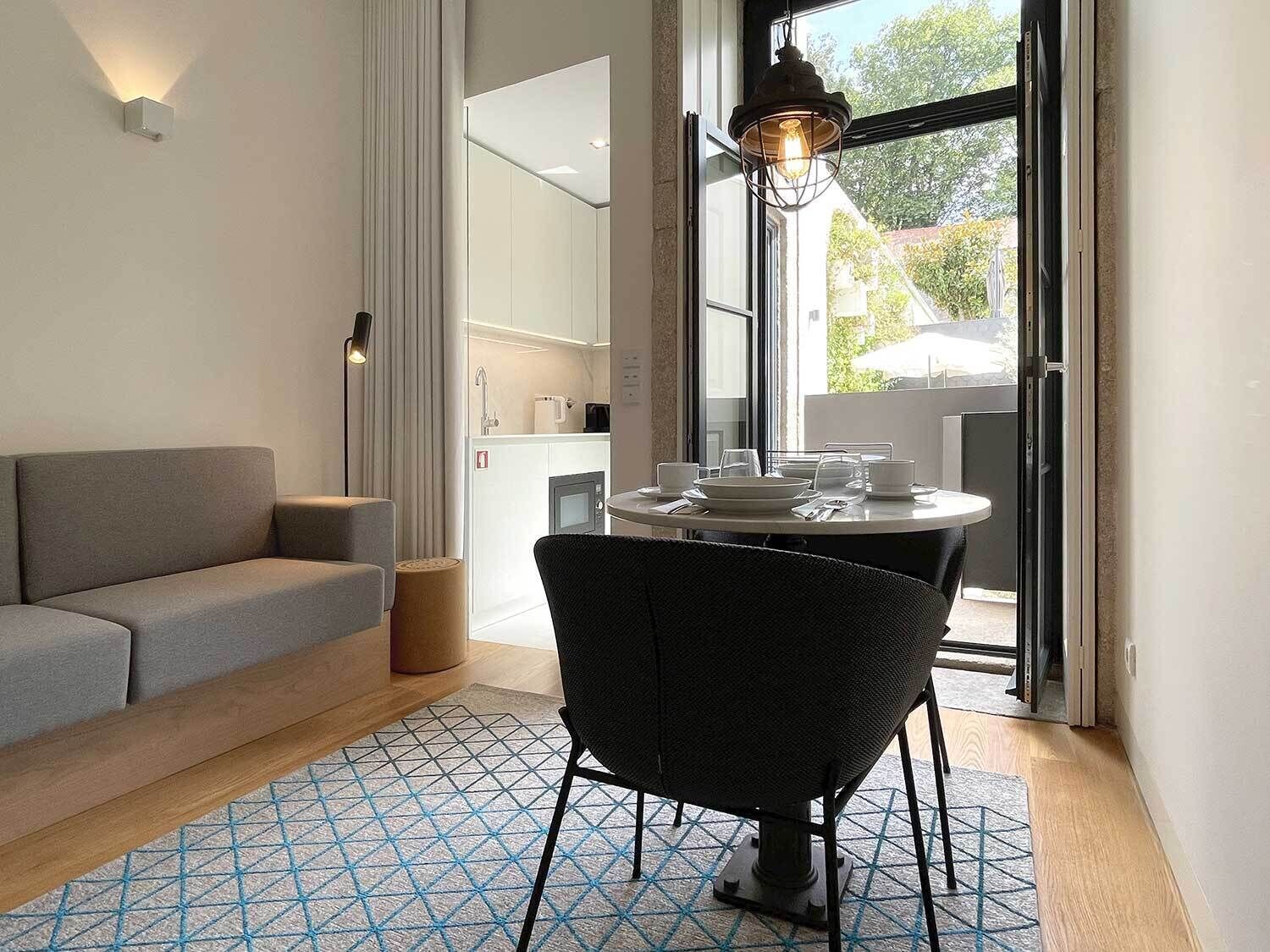 A modern living room with a sofa, a dining table with chairs, a patterned rug, and a kitchen area visible through an open door.