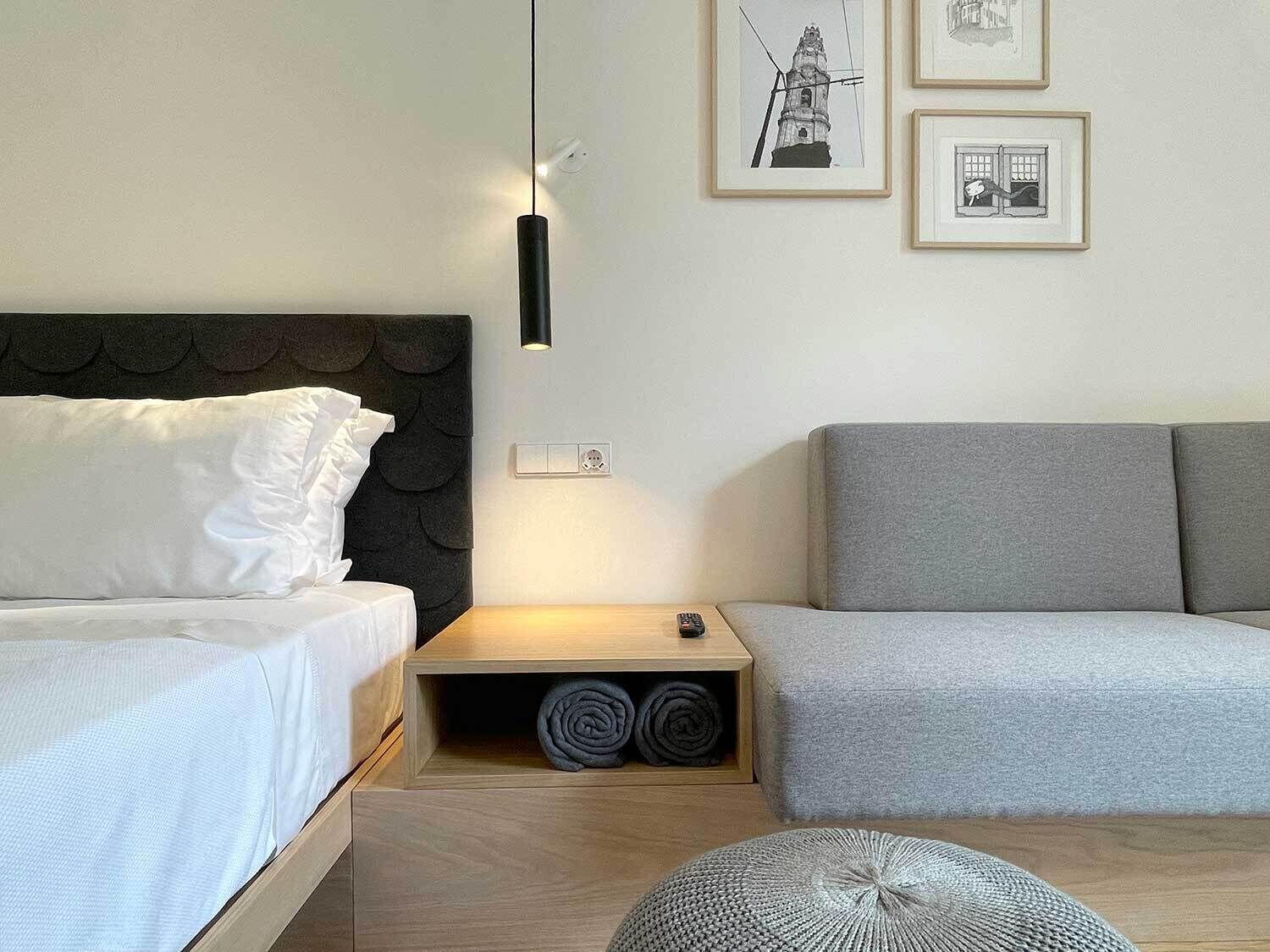 Un dormitorio moderno con cama, un sofá gris, una mesita con lámpara y una pared con cuadros enmarcados.