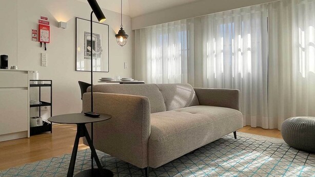 A modern living room with a sofa, a small table, a lamp, and a large window with sheer curtains. - Essência Portuense Studios (Porto)