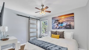Quarto com cama, escrivaninha, ventoinha no teto, janela e um quadro na parede.