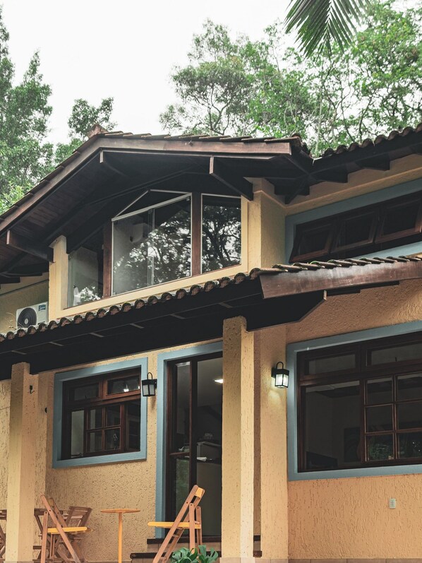 Un edificio de dos pisos con balcón, ventanas amplias y un patio cubierto con mobiliario de madera.