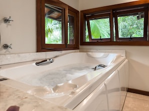 Un baño con una bañera grande, marcos de ventana de madera y vistas a la vegetación exterior.