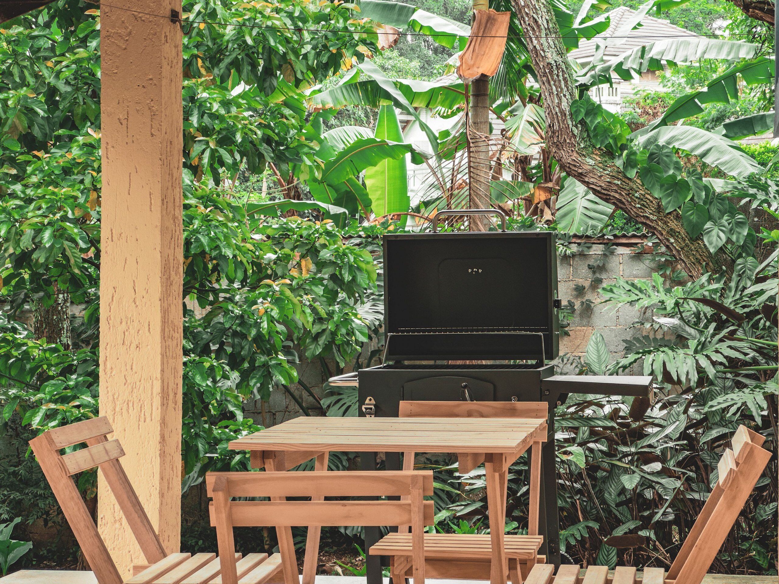 Une table et des chaises en bois, installées à l’extérieur près d’un barbecue noir, sont entourées d’un luxuriant feuillage verdoyant.