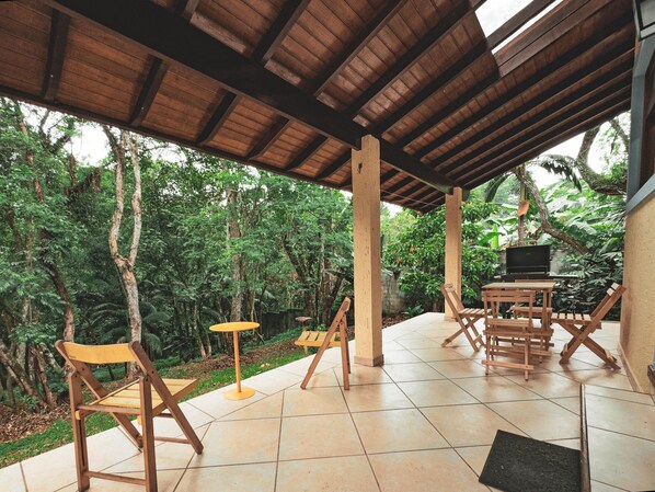 Un patio cubierto al aire libre con mobiliario de madera, una parrilla y abundante vegetación en el fondo.