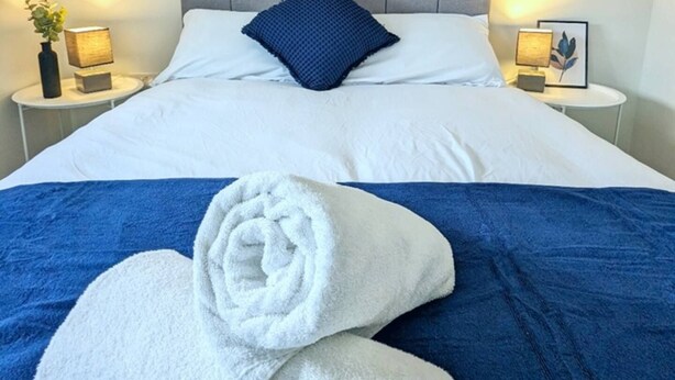 Een opgemaakt bed met witte lakens, een blauw sierkussen en twee opgerolde handdoeken op een blauw sprei.