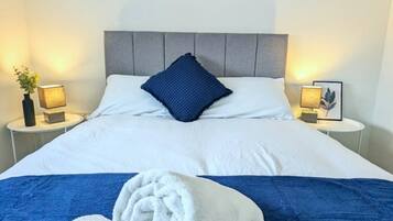 A neatly made bed with white linens, a blue accent pillow, and two rolled towels on a blue bedspread.