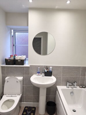 Une salle de bain avec des toilettes, un lavabo et une baignoire. Il y a un miroir au-dessus du lavabo et une fenêtre donnant sur l’extérieur.