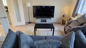 Een woonkamer met een bank, een televisie op een standaard, een vloerlamp en een raam met gordijnen.