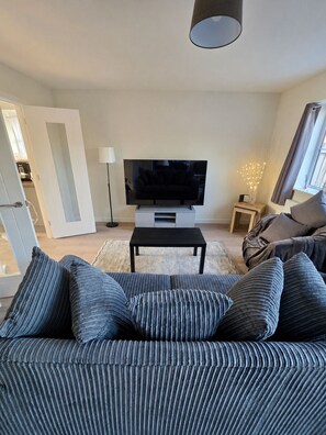 Un salon avec un canapé, un téléviseur posé sur un meuble, une lampe sur pied et une fenêtre avec des rideaux.