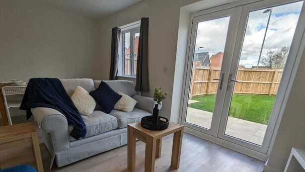 A living room with a grey sofa, a blue armchair, a wooden coffee table, and a sliding glass door leading to a backyard. - Watton House | 3 bedroom house with garden | Ipswich, London (Carbrooke)