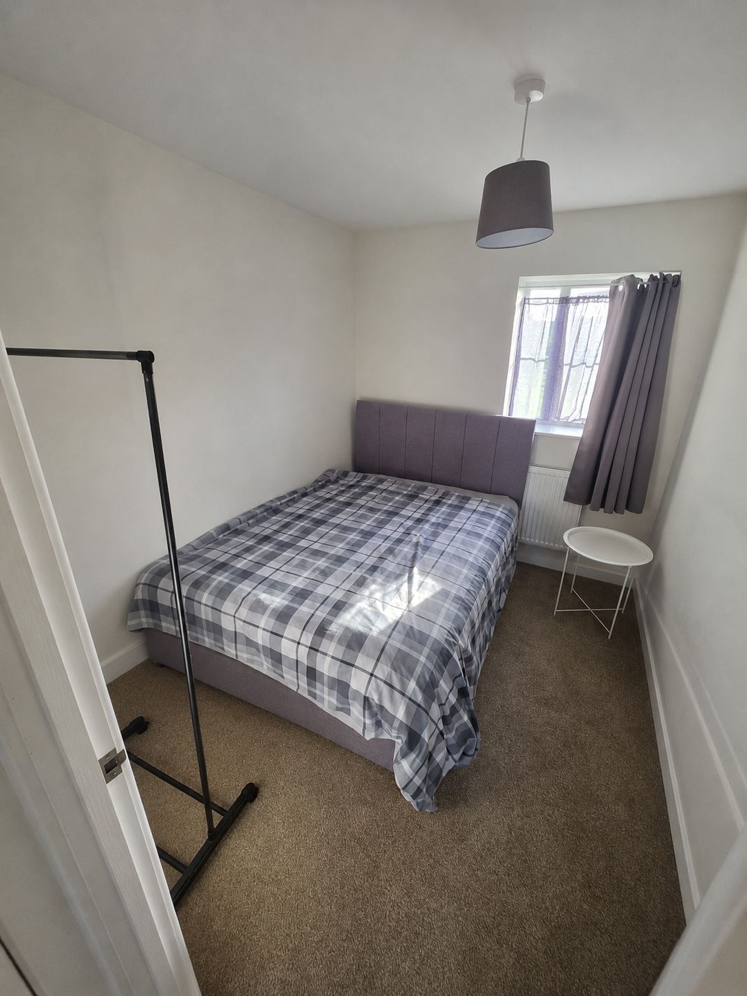 Une chambre avec un lit, un ventilateur sur pied, une fenêtre avec des rideaux et une petite table.