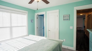 Une chambre à coucher comprenant un lit, un ventilateur de plafond, une fenêtre avec des stores et une porte donnant sur une autre pièce.