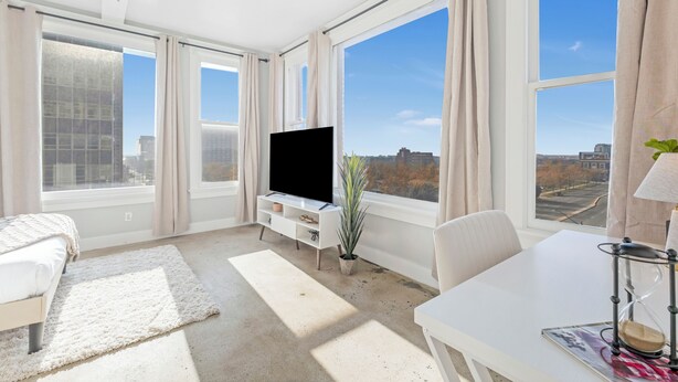 A modern hotel room with large windows, a flat-screen TV, a dining table, and a cozy seating area.