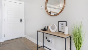 A hallway with a white door, a round mirror, a framed welcome sign, a small table with a router, a vase, and a potted plant.