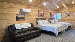 A cozy room with wooden walls and ceiling, a leather sofa, two beds with patterned bedding, and a mounted air conditioner.