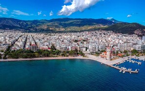 An aerial view of a coastal city with dense urban development, a prominent church with a bell tower, and a marina with boats. - Fili's Central Spots (Volos)