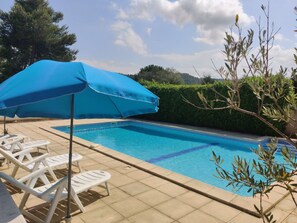 Une piscine avec un parasol bleu, des chaises longues blanches et une haie en arrière-plan.