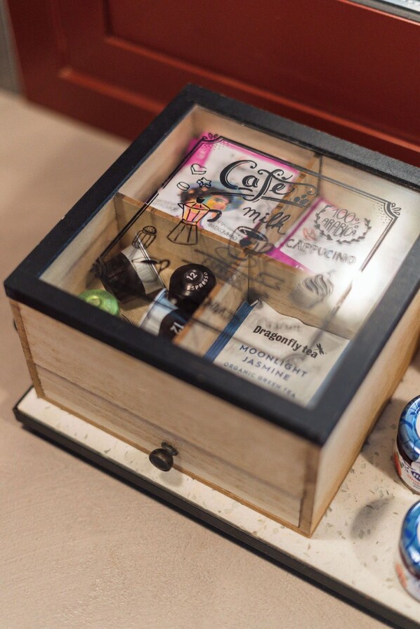 Eine Holzkiste mit Glasfront, die verschiedene Gegenstände beherbergt, darunter eine Kaffeemaschine, ein Teebeutel und eine Cola.