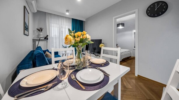 A dining area with a white table set for four, a vase of yellow flowers, and a clock on the wall. - Warsaw Airport Apt- Parking&Terrace - by Rentujemy (Warszawa)