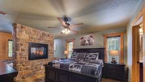 A bedroom with a stone fireplace, a ceiling fan, a bed with a patterned quilt, and a nightstand with a lamp. - Luna Bear Lodge (Gatlinburg)