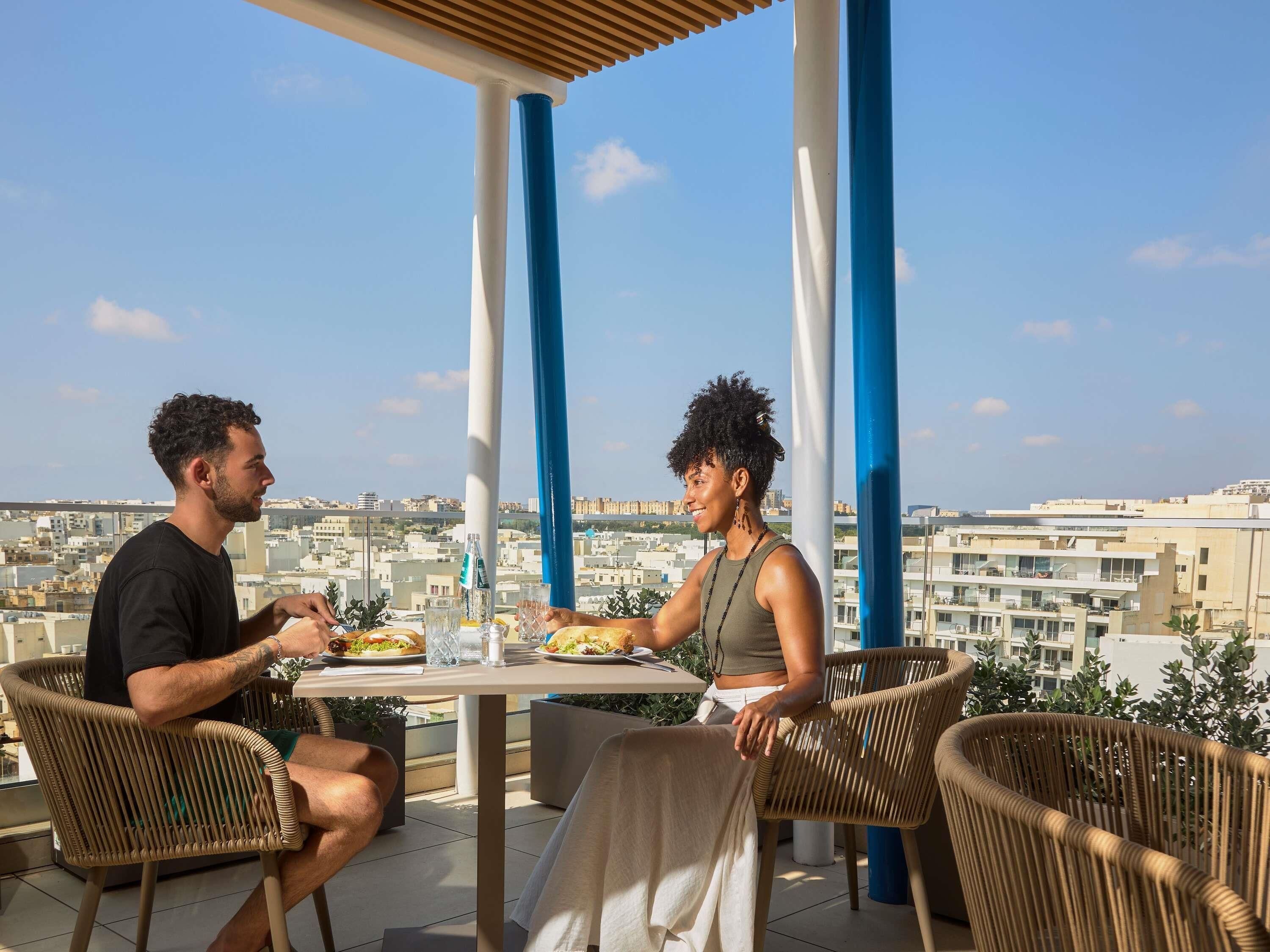 Two people dining at an outdoor table with a city view, under a covered area with blue and white pillars.